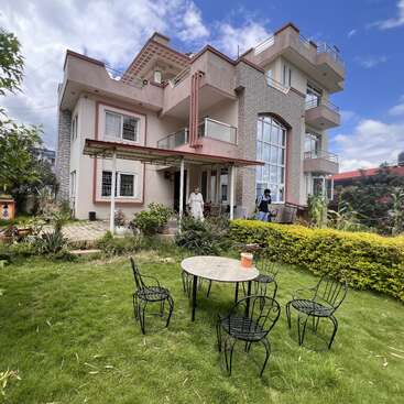 A modern two-story house with balconies stands amid a lush green garden, featuring a round table with four chairs, under a partly cloudy blue sky.