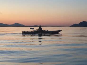 The image depicts a serene scene of a person kayaking on a tranquil body of water at sunset, with mountains visible in the distance.