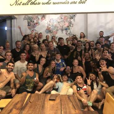 The image depicts a group of approximately 40 people posing for a photo, with a wooden table in the foreground and a world map on the wall behind them.