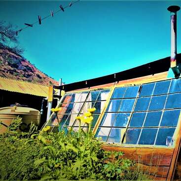 The image depicts a rustic wooden building with solar panels on its side, featuring a window and a chimney, set against a hillside and clear blue sky.