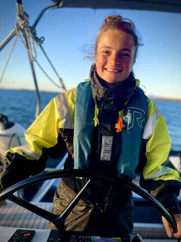 The image depicts a woman wearing a life jacket and standing at the helm of a boat on a body of water, with a clear blue sky in the background.