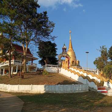A golden pagoda stands atop a hill, surrounded by trees and a white fence. A staircase adorned with golden serpents leads up to the shrine.