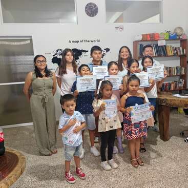 A group of children and adults pose indoors, proudly holding certificates. They stand in front of a world map wall, bookshelves, and smiling for the camera.