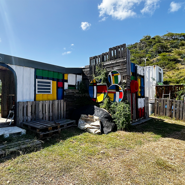 The image depicts a vibrant outdoor space with a shipping container repurposed into a colorful structure, surrounded by lush greenery and a serene blue sky.