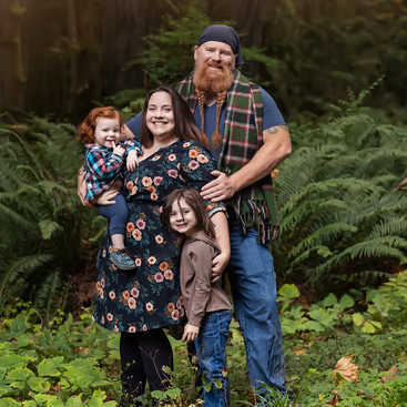 A happy family of four poses outdoors, surrounded by lush green ferns and plants. They are smiling, dressed warmly, and radiate warmth and togetherness in nature.