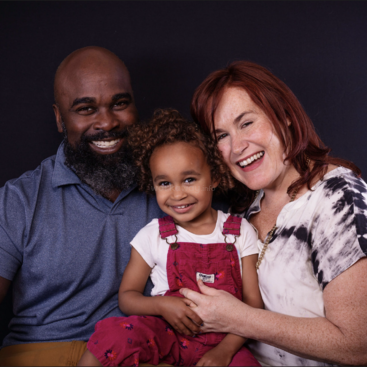 The image depicts a family of three posing for a photo, with the father on the left, the daughter in the middle, and the mother on the right, all smiling at the camera.